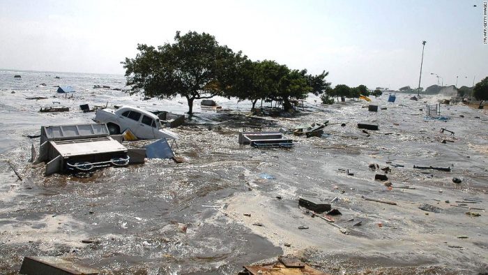 Flood waters wash over trees, debris, and cars.